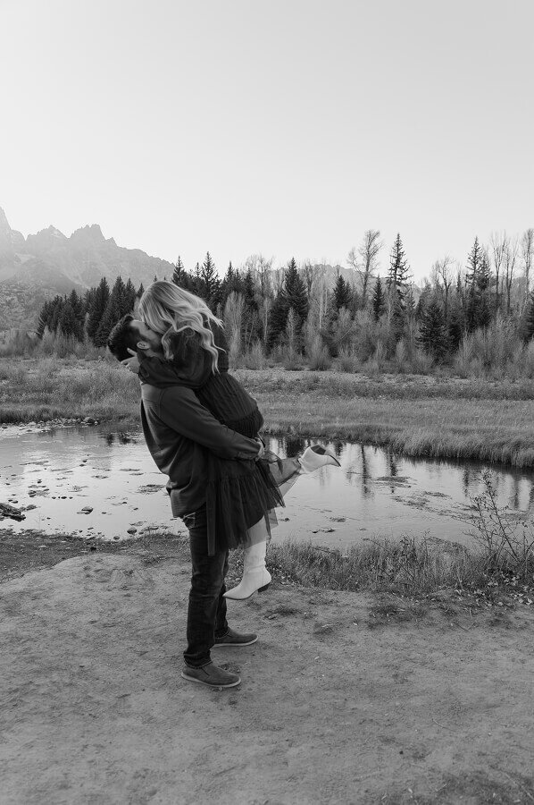 A black and white photo in Grand Teton National Park of a newly engaged couple. He is picking up his fiancé off the ground and they're kissing so you don't see their faces.
