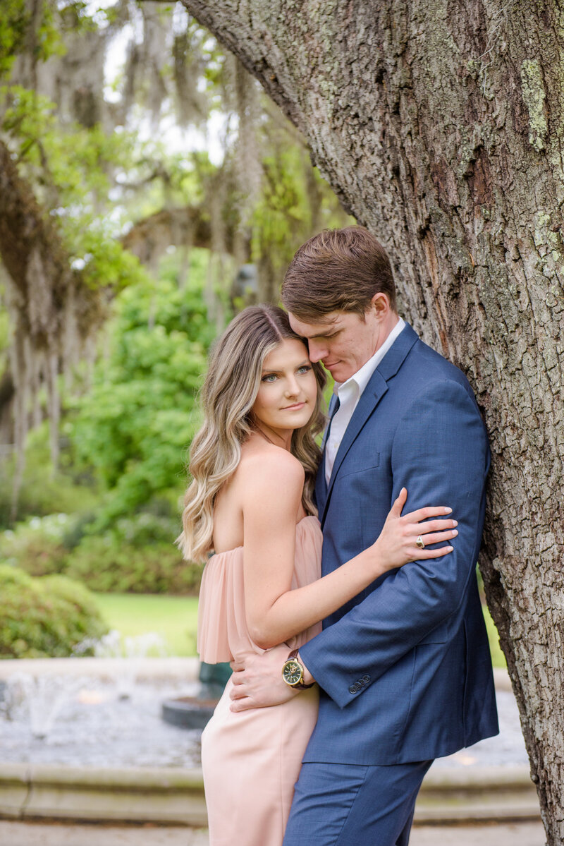 Couple shares a kiss during their engagement session beneath large oak trees, with the bride-to-be in a short white dress and the groom-to-be in casual attire.