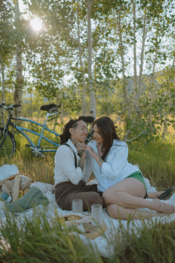 A woman and her fiancé sit on a picnic blanket smiling at each other for their engagement couples shoot.