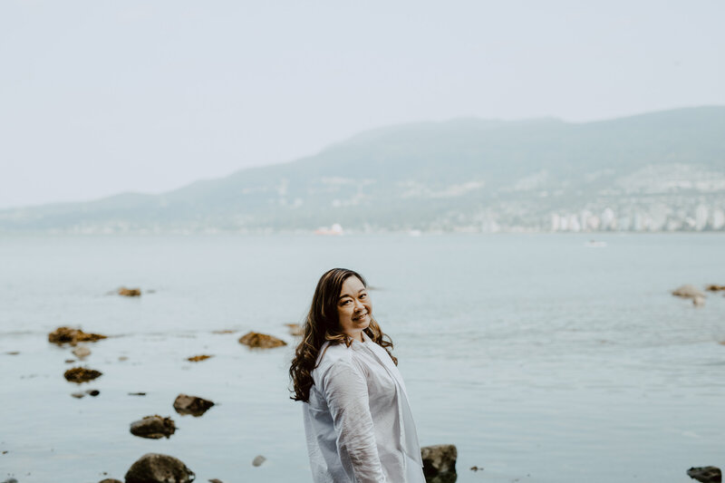 Anne, an Asian woman, is looking over her shoulder at the camera and smiling. She is wearing a white dress. In the background is a view of the North Shore Mountains with water view .
