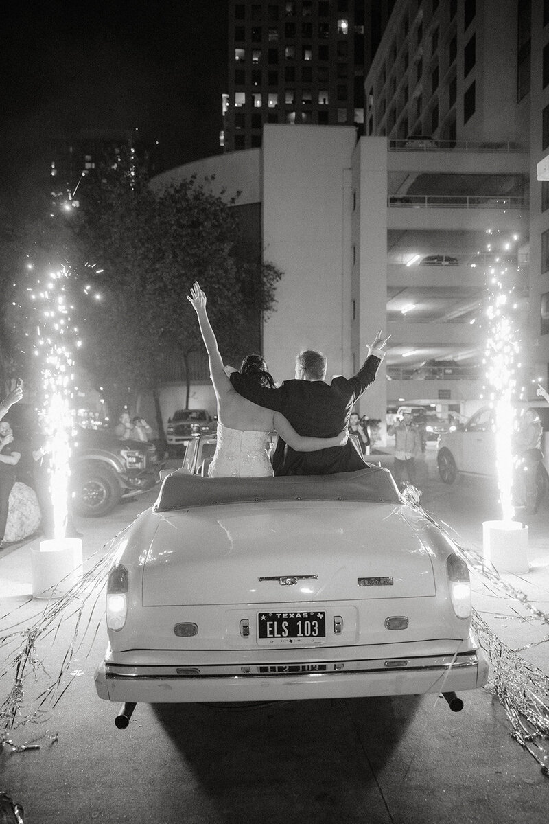 Black and white photo from behind of bride and groom sitting on the back of their wedding getaway car with cold sparks around them