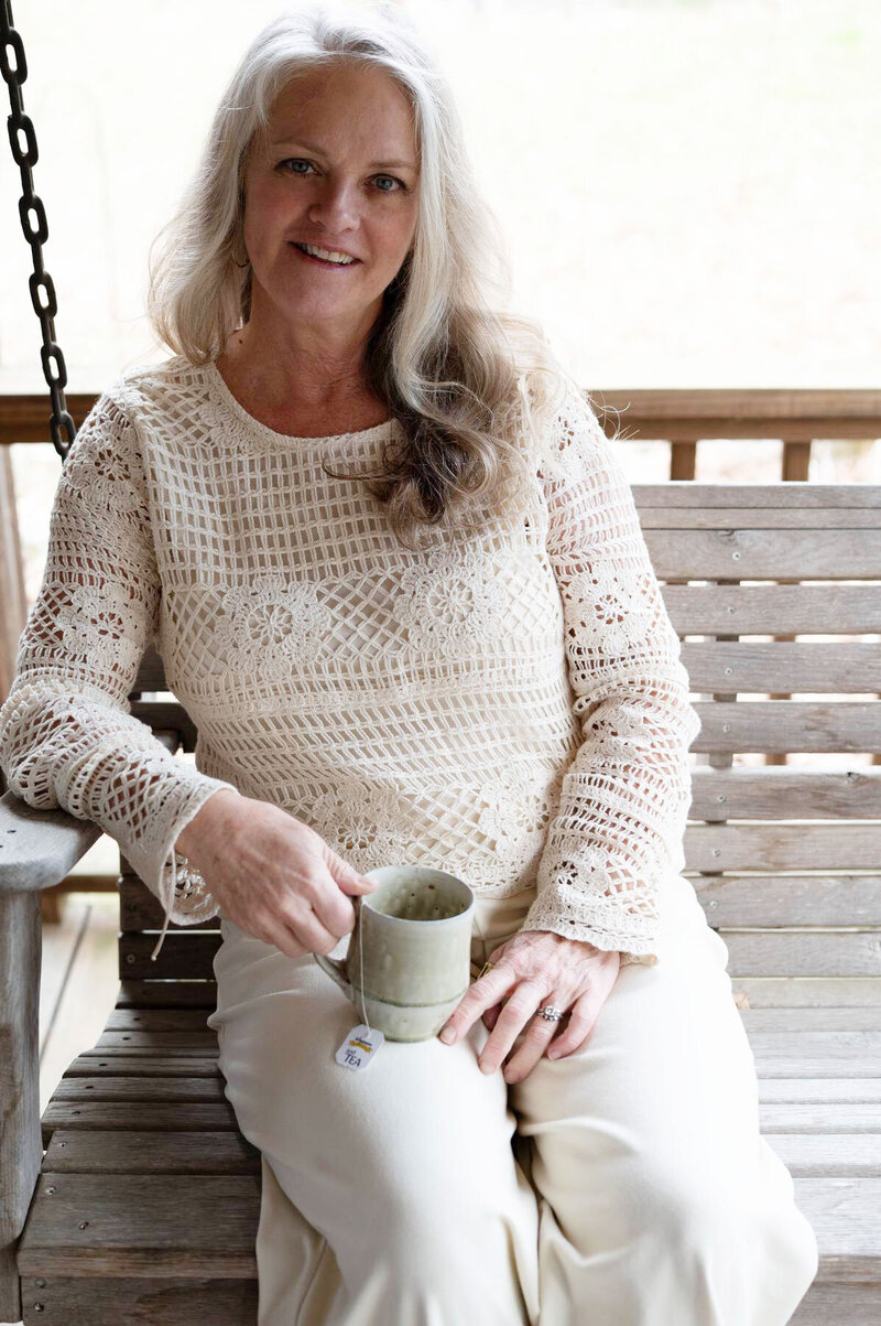 Janna Weldum of Eagle's Guiding Light sits on a porch swing holding a tea cup with a warm, welcoming smile on her face.