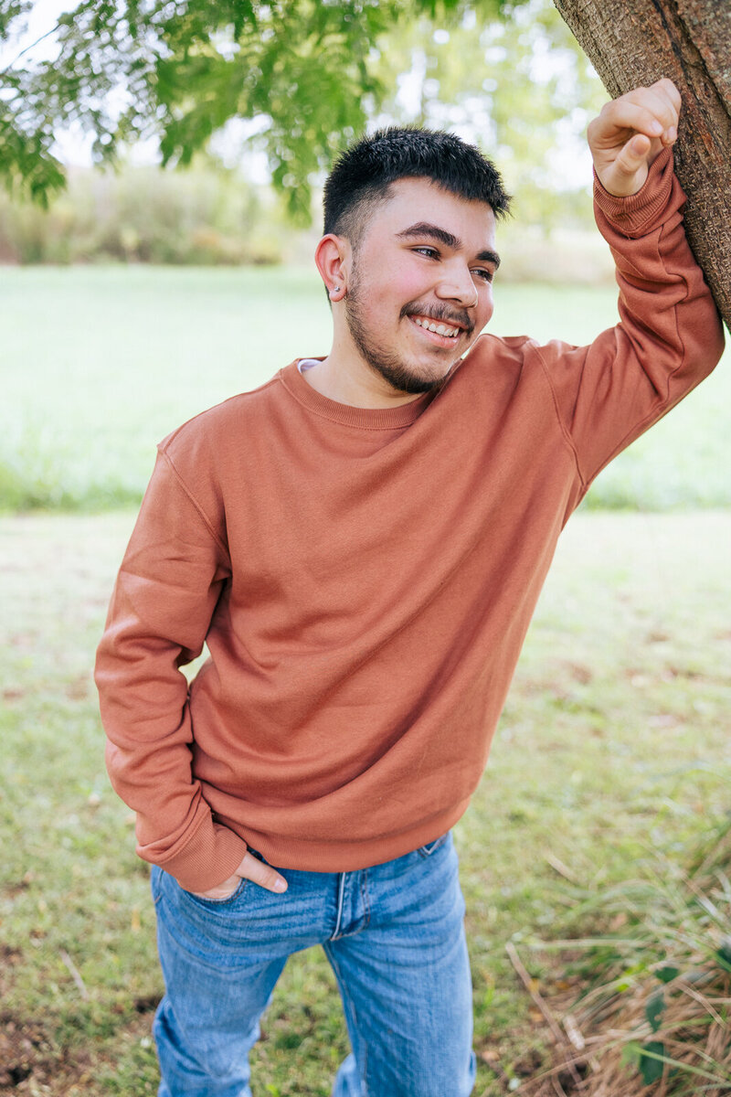 Senior guy leaning on tree with his hand in his pocket smiling. 