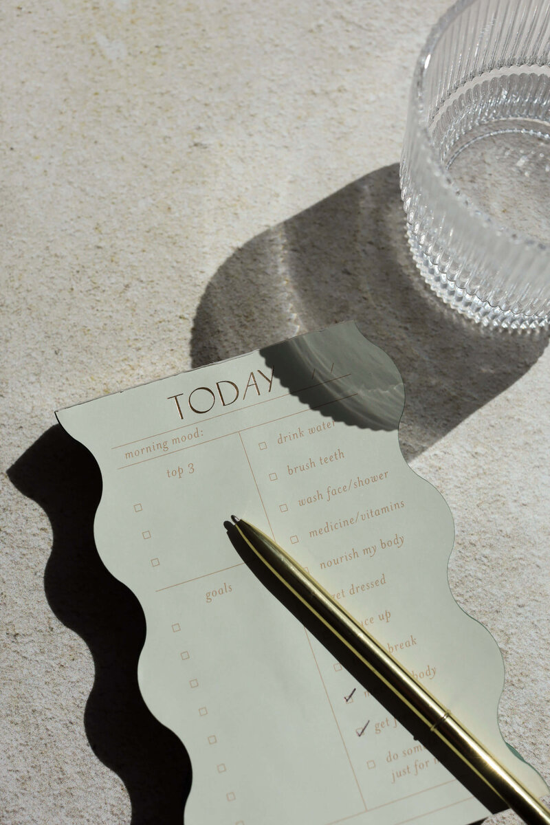 A notepad with a pen on a table, accompanied by a glass of water, reflects a peaceful environment for organizing self-care priorities.