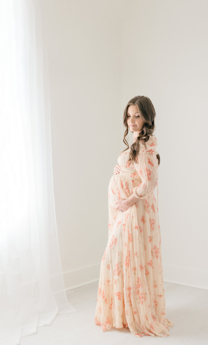 Pregnant woman standing next to window in an all white studio by NJ Maternity Photographer