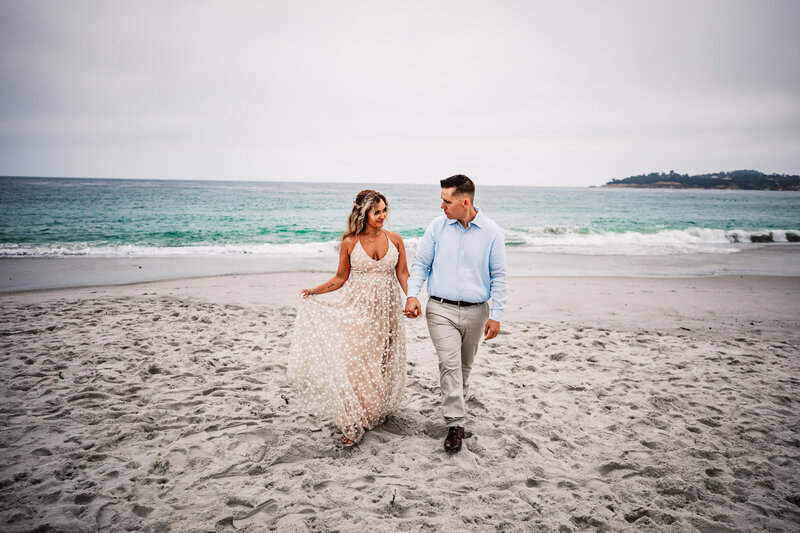 A couple lovingly walks hand-in-hand along a sandy beach, smiling warmly at each other. The bride wears a flowing, blush-colored dress, and the groom wears a light blue shirt and beige pants. Gentle ocean waves break softly behind them under an overcast sky.