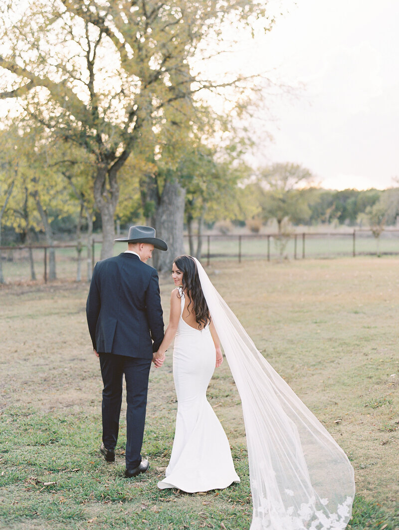 Bride looking back at the camera while walking and holding hands with groom in a cowboy hat