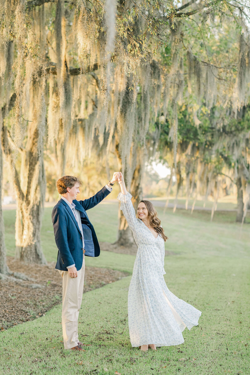 Man holding womans hand under glowy trees while she dances for engagement phtoos
