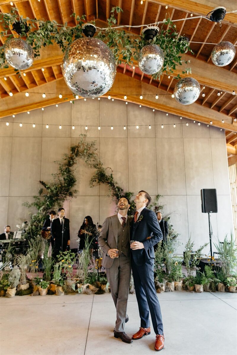 Two grooms admire the elegant disco ball and botanical decor alongside twinkle lights at industrial modern Catskills wedding venue