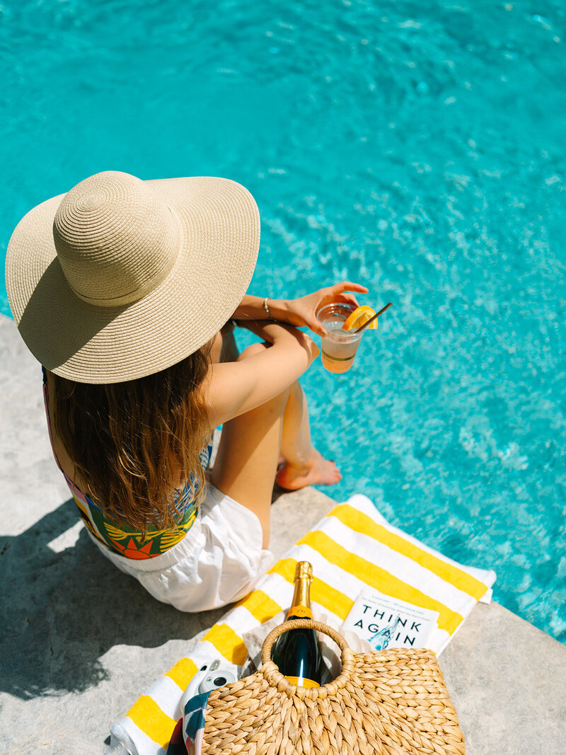 Woman in a straw hat sitting on a yellow striped towel with a drink beside a pool