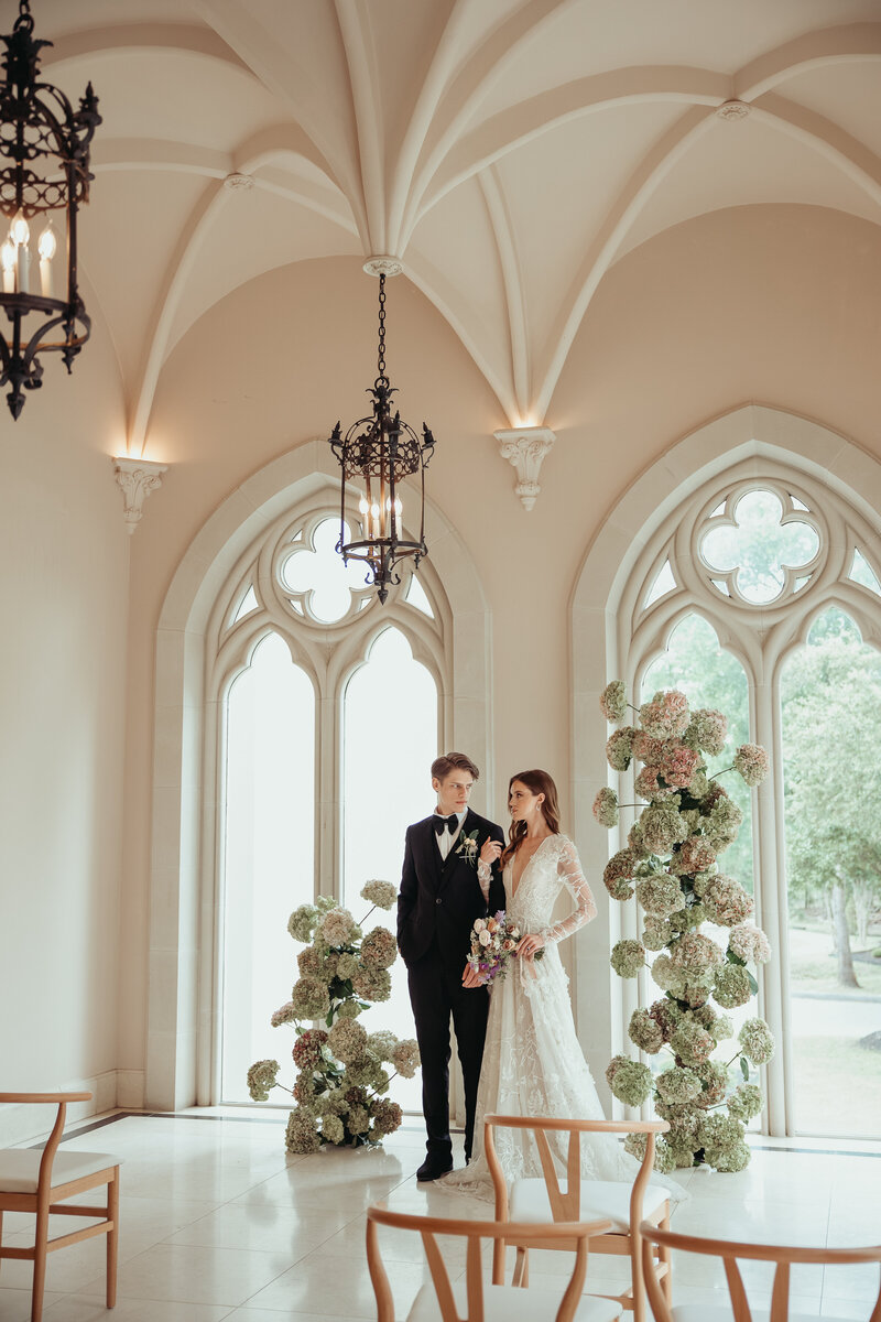 Couple standing in a light-filled room with gothic arched windows, sculptural floral pillars, and minimal ceremony seating.
