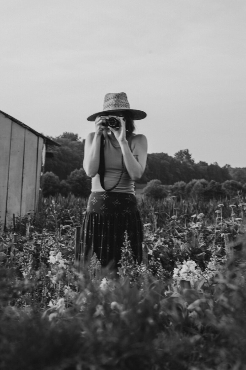 A person standing in a field of tall flowers as they hold a camera up to their face 
