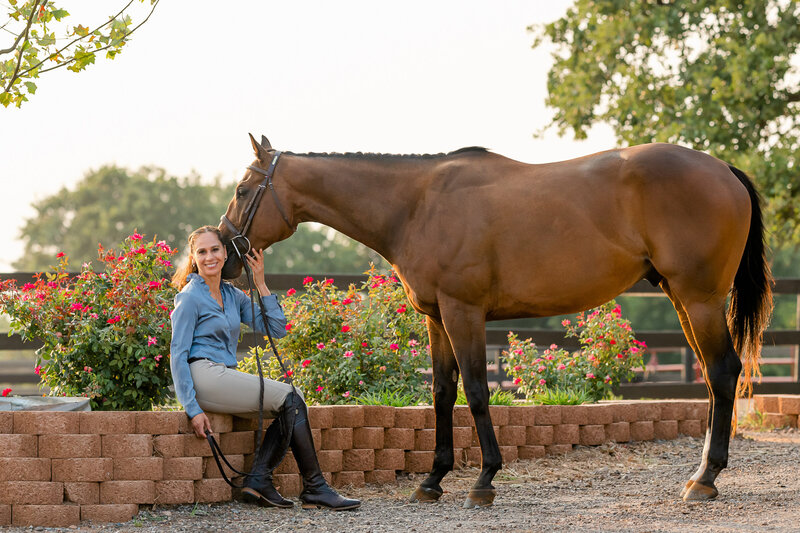 An adult amateur equestrian sits near roses and poses for portraits with her Oklahoma Bred Off The Track Thoroughbred Phelps during mother daughter horse portraits at the woodlands in Edmond Oklahoma
