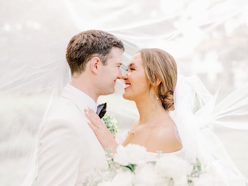Male and female couple poses for photographer smiling at each other, holding blush and white flowers and wearing a long white veil at their Dallas wedding.