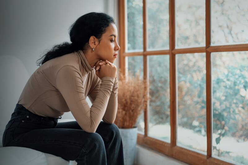  A woman with dark hair tied back in a ponytail sits sideways on a white couch, leaning forward and gazing out a large window with her hands folded under her chin.