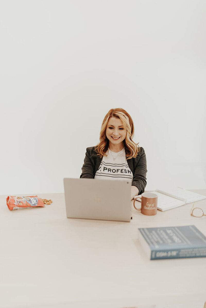 Shylah sits at a large desk, smiling down at her laptop, with a coffee mug, notebook, glasses, and a bag of Nerds candy surrounding it on the desk. She is wearing a t-shirt that says "V. PROFESH", with a blazer over top of it.
