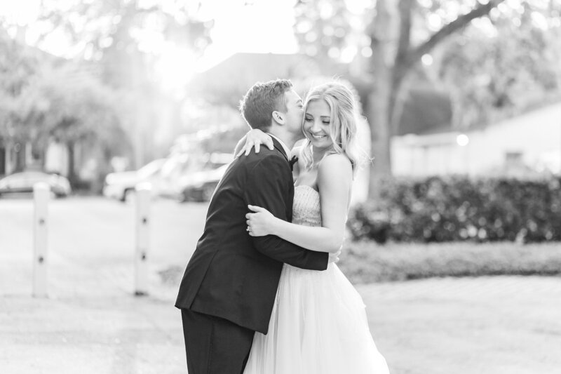 Bride smiling while groom kisses her cheek in black and white during their wedding at The Orlo