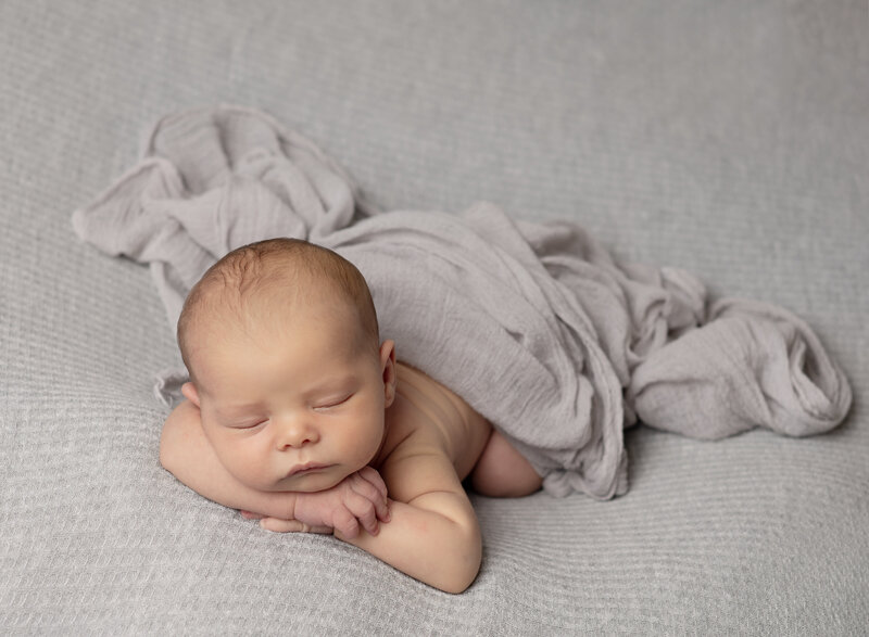 newborn boy laying on his belly resting his chin on his hands on light gray backdrop