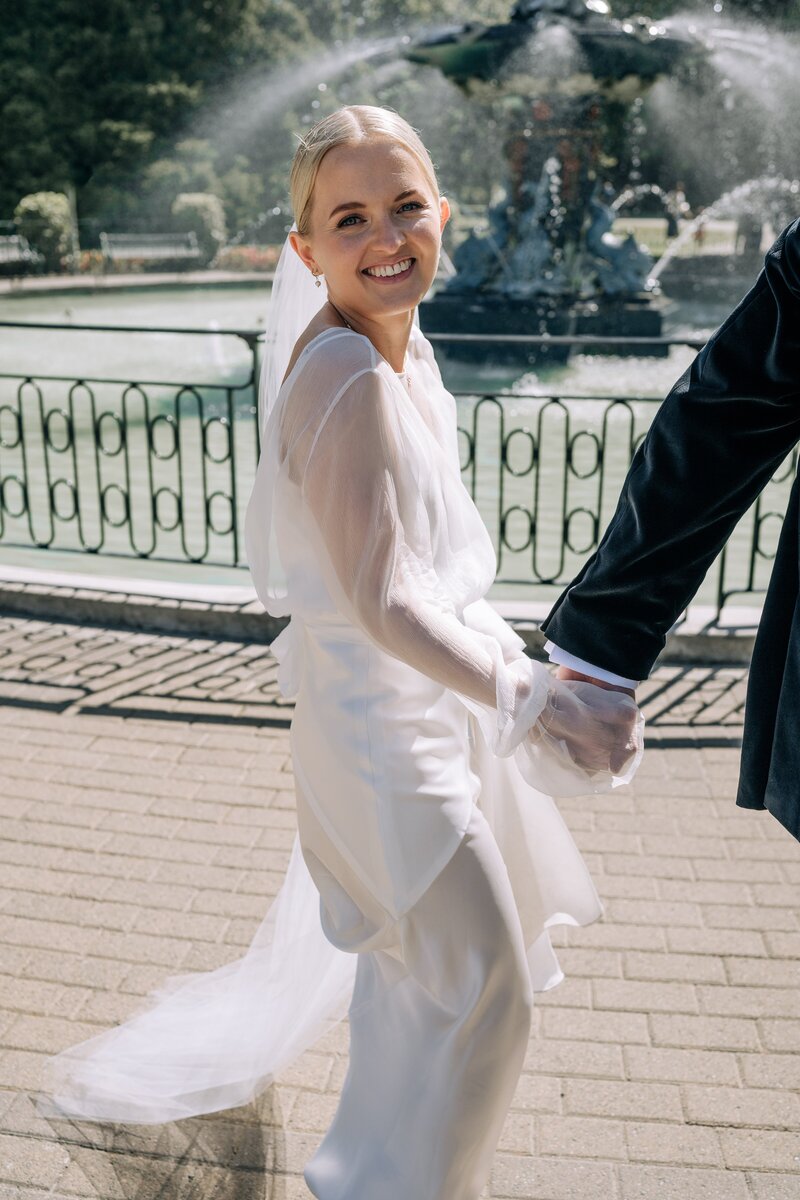 candid photo of a bride smiling as she walks past the fountain in the christchurch botanic gardens on her wedding day