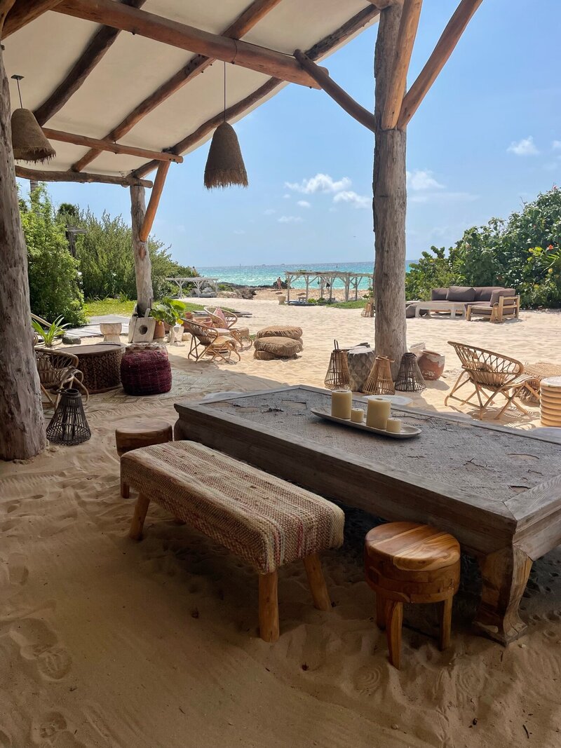 A rustic, open-air dining area on a sandy beach, with wooden benches, tables, and woven seats, set up with a view of the ocean in the background. The peaceful setting is enhanced by a few decorative candles on the table.
