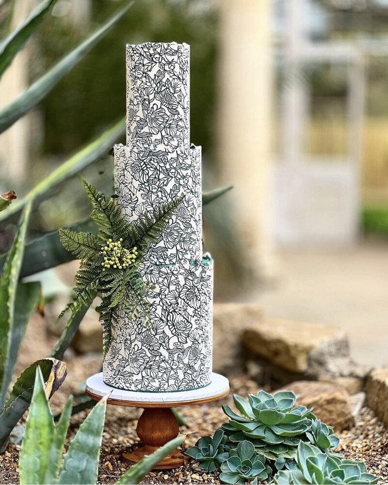 Three tier wedding cake in emerald green & white  in stencilled buttercream with a cluster of natural green foliage on wooden cake stand