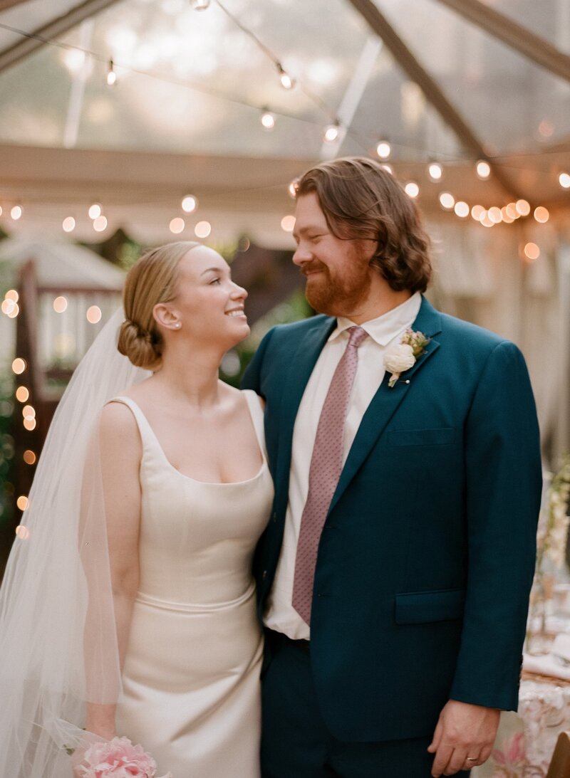 Soft film portrait of a bride and groom smiling at each other under string lights on their wedding day — captured by a film wedding photographer at Castillo Holliday Photo + Film.