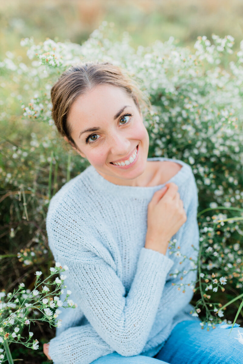 Portrait of a girl sitting in white and green wildflowers. Her hair is in a low bun and she is wearing a knitted grey sweater.