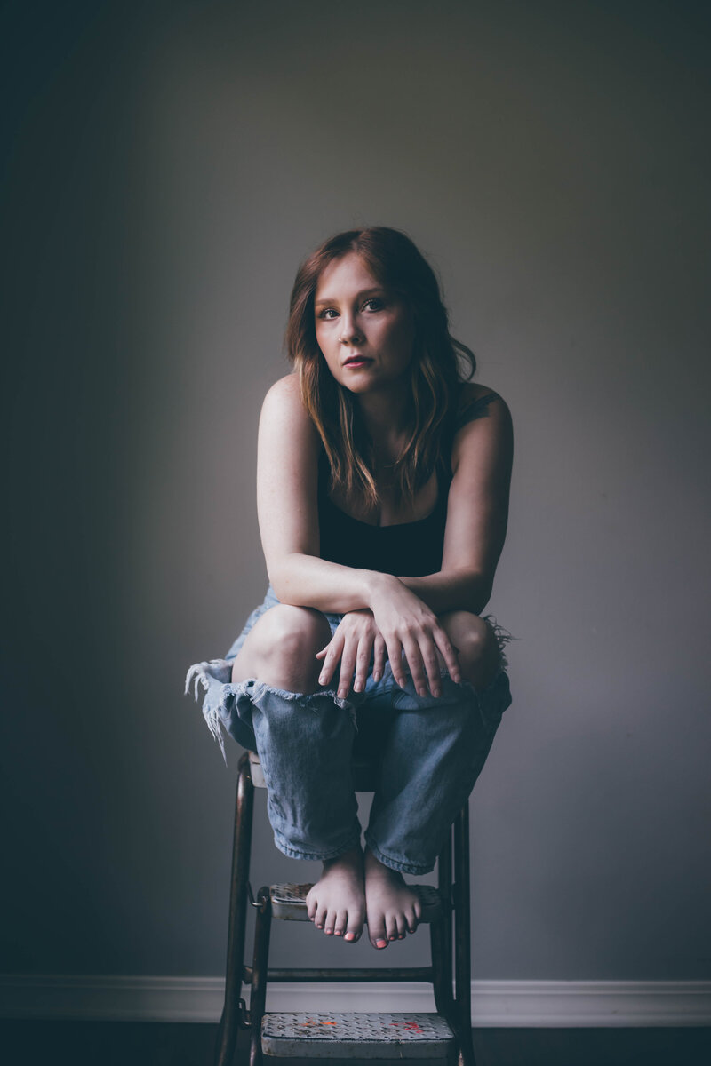 A person with long hair, dressed in a black tank top and ripped jeans, sits on a stool against a plain background, looking forward—ready to create your website in a day.