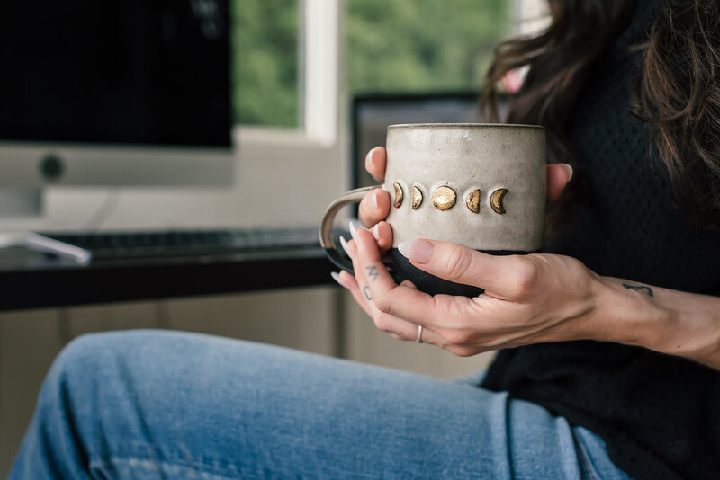 Miranda holding a ceramic mug adorned with golden moon phases, sitting at her desk in a peaceful workspace.