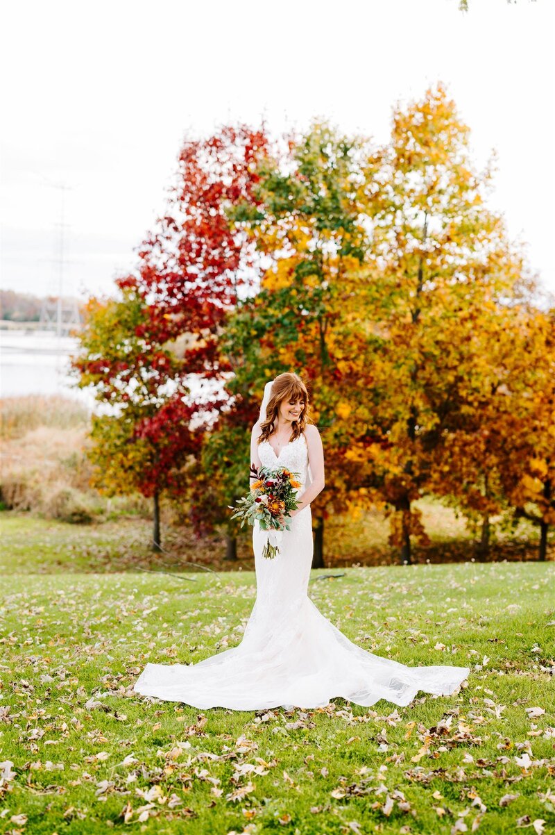 A bride in a long, elegant white wedding gown stands on a grassy lawn covered with fallen leaves. She holds a bouquet of autumn flowers and looks down with a gentle smile. Behind her is a stunning backdrop of trees in peak fall colors, with leaves in shades of red, yellow, and orange, near a serene lake.