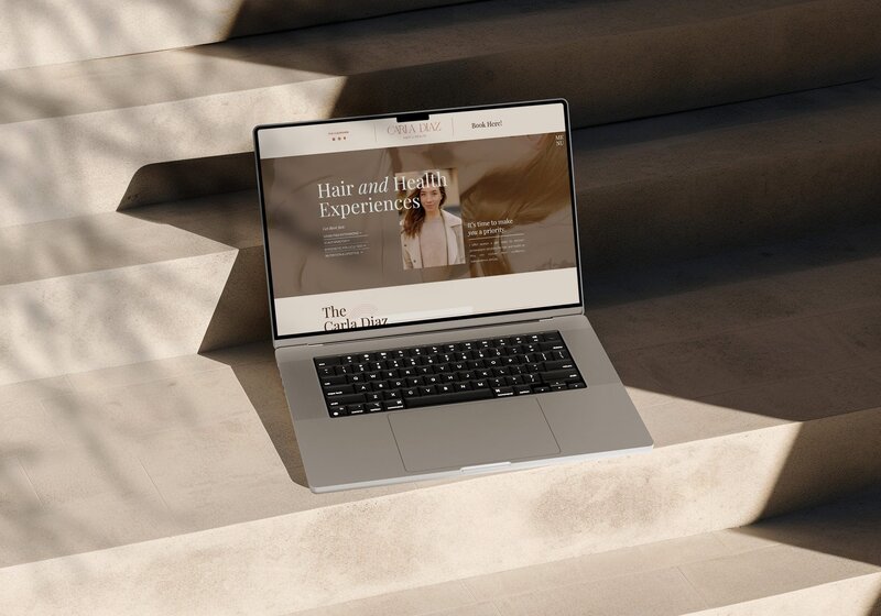 A laptop displaying a website with the headline "Hair and Health Experiences" rests on concrete steps. Shadows are cast over the scene, highlighting the sleek logo and web design tailored for salons.