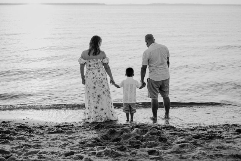 Black and white photo or mom, toddler, and dad standing on the beach in Kent Island Maryland looking at the bay.