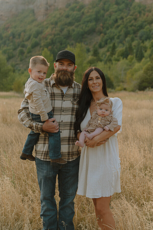 Holding their young baby girl, a mother stands next to her husband whose holding their toddler son. They're smiling for a family photo.