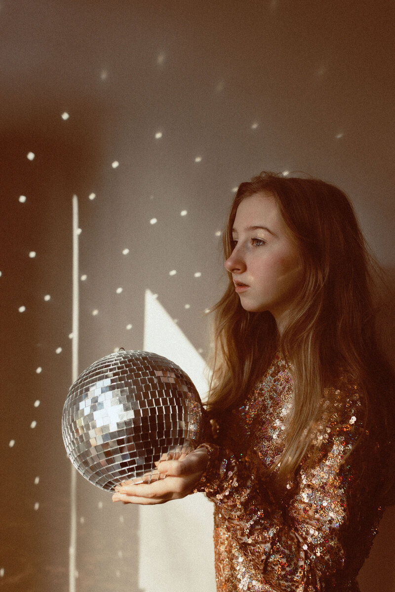 A senior girl holding a disco ball that is reflecting the light onto the wall in a magical way. 