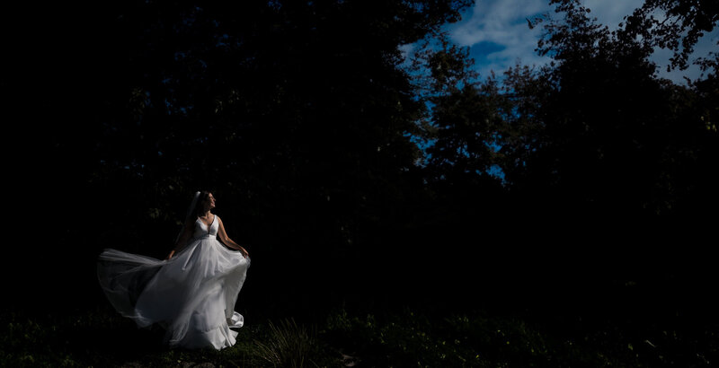 Braut in einer dramatischen Pose vor einem dunklen Hintergrund, ihr Kleid weht im Wind und sie schaut in die Sonne