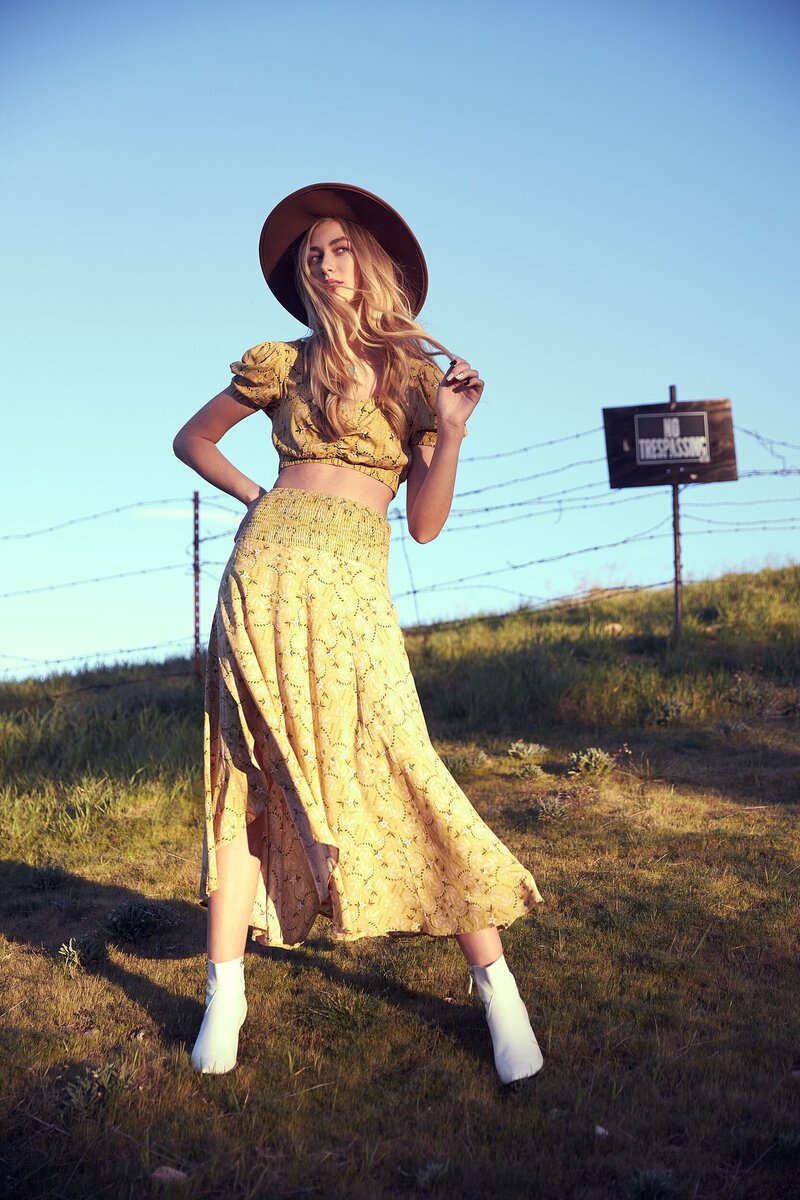 Editorial senior photo of a stylish teen girl in a yellow boho crop top and skirt, wearing a wide-brim hat and posing boldly in a sunlit field with a No Trespassing sign in the background.