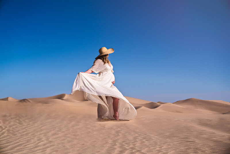 Mom standing in desert against sandy hills and blue skies. Mom is standing side profile to the camera wearing a long, flowing dress and wide brimmed hat. Her back arm is cradling her bump and her forearm has her dress extended behind her and flowing with the wind. Captured by top Philadelphia and Mainline maternity photographer Liza Nicole