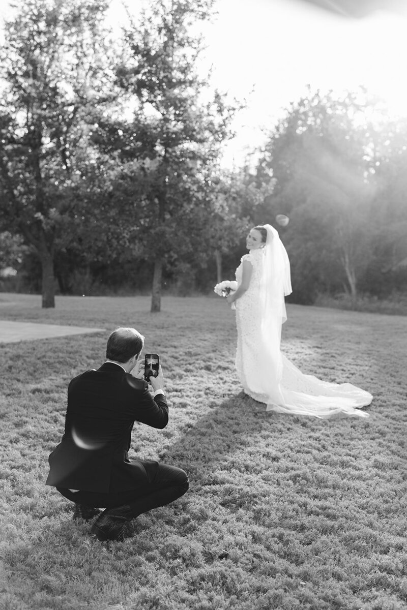 Black and white photo of groom taking iphone photo of his new bride at the wedding venue