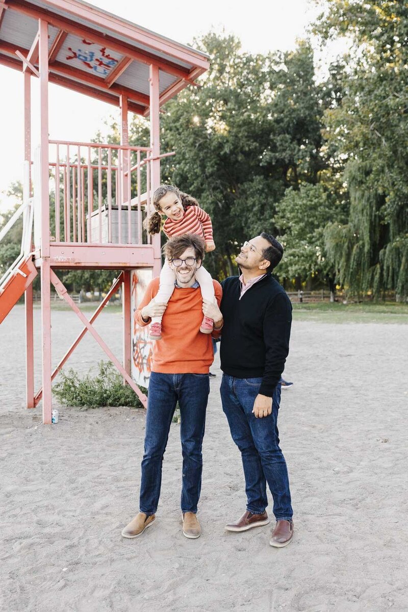 Two people stand on a sandy area near a orange lifeguard stand during their Toronto family portrait session. One carries a child on their shoulders, wearing a charming red and white striped shirt. Trees create a picturesque backdrop in this serene moment.