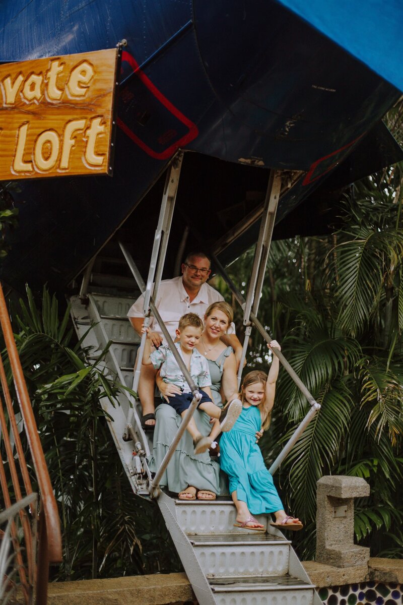 Family of six sitting together on loft stairs at a tropical loft-style venue, surrounded by lush greenery.