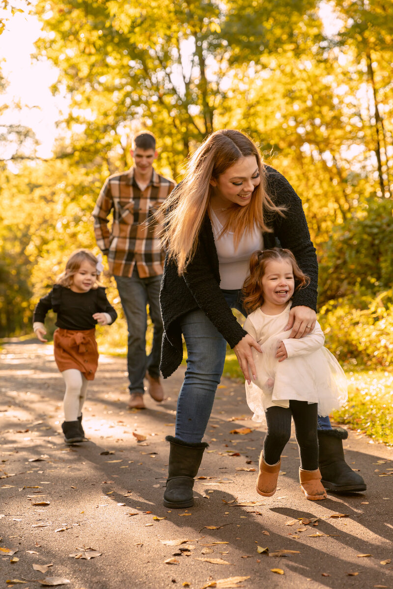 young family of four with two girls chase each other on a path through the yellow fall woods at Parker Mill County Park in Ann Arbor michigan