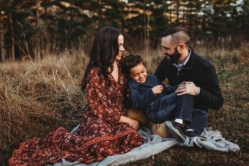 fall family pictures with Baltimore photographers capturing mother in a red dress sitting with her son and husband on a blanket with the woods in the distance