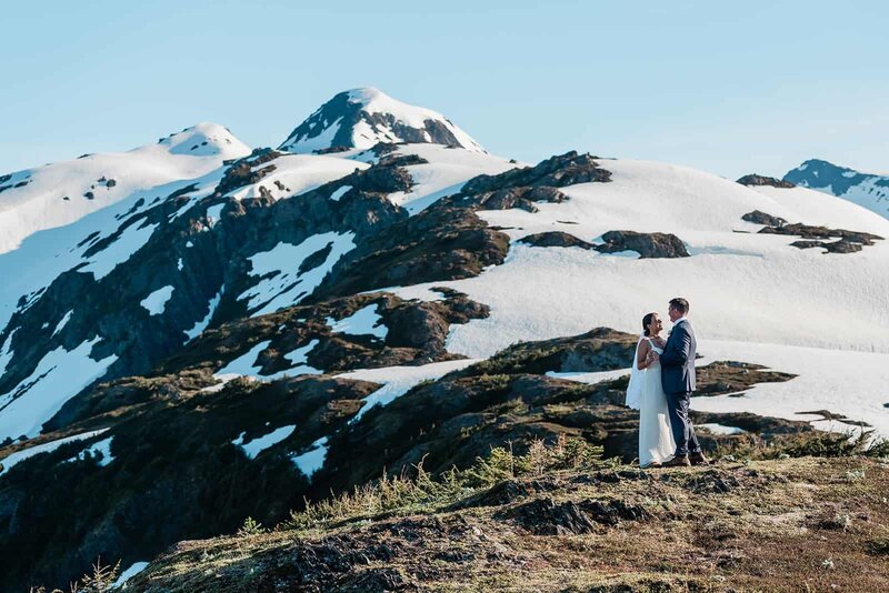 A couple in their wedding finest does their first dance on the edge of a mountain top during their private helicopter elopement package in Seward Alaska.  The rock and tundra surrounds them.  Behind them are craggy mountain peaks, with spots of snow interspersed between rock outcroppings.  The sky is bright blue.   