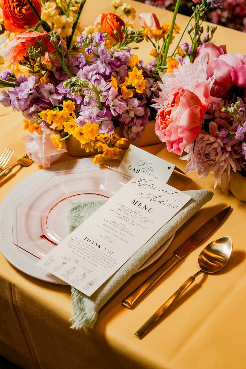 Bright florals on a yellow tablecloth with gold settings, a menu, and a porcelain plate.
