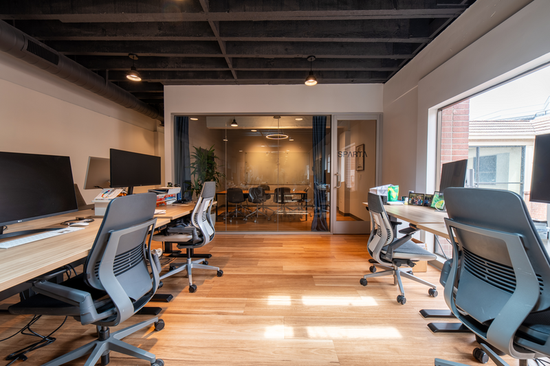 Modern office with long wooden desks, ergonomic chairs, and a glass-walled conference room labeled Sparta.