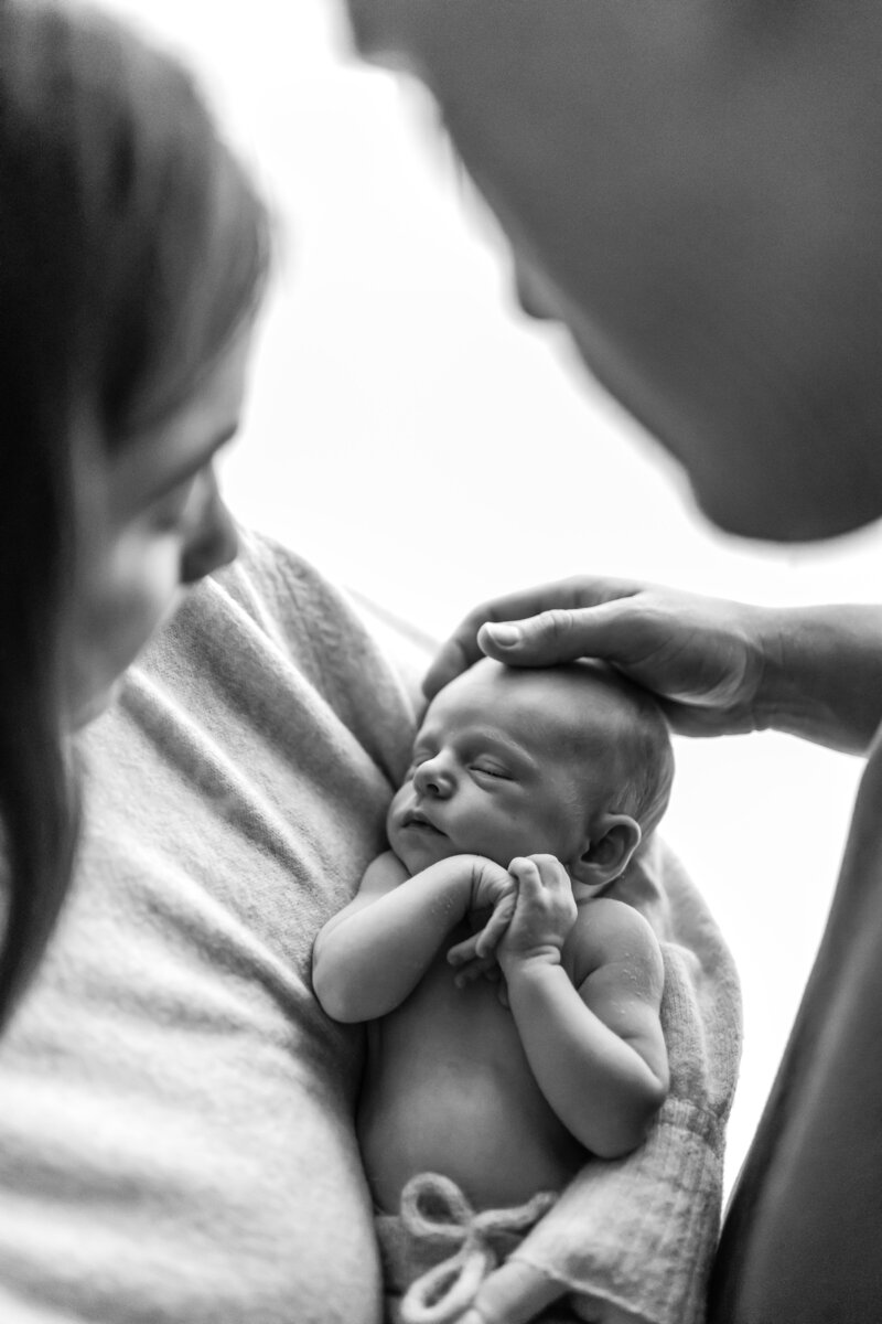 Mother holds a sleeping newborn while dad rests his hand gently on their head black and white image
