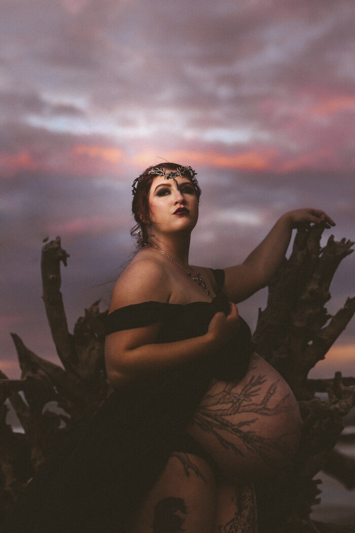 A maternity portrait of a woman in a black gown and crown at sunset, seated on driftwood under a dramatic purple sky.