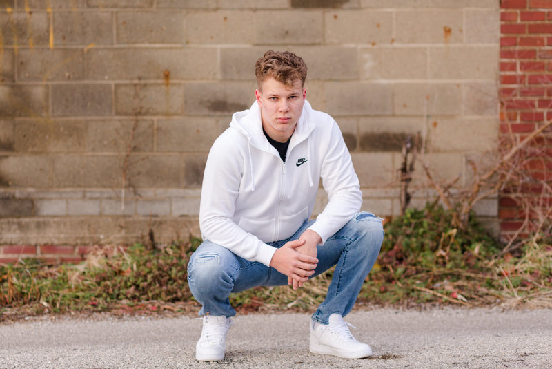 waynedale high school senior guy wearing blue long sleeve shirt, with tan pants, sitting in fwaynedale high school senior guy squatting in an alley way wearing nike shirt and nike shoes with jeans, serious look on his face,, photographed in Wooster Ohio, photographed by Jamie Lynette Photography Canton Ohio Senior Photographer