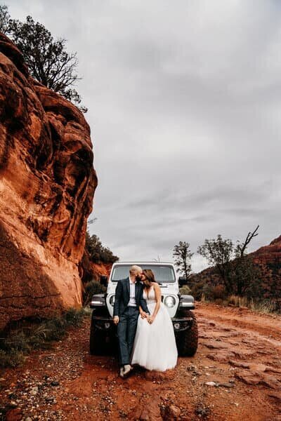Bride and groom cuddling in front of jeep headlights in their wedding attire while eloping in Sedona Arizona on the trail to Merry Go Round Rock with their elopement photographer