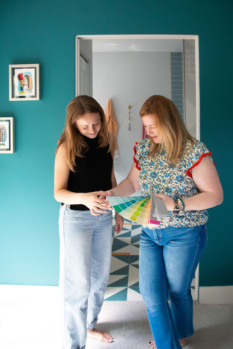Two women are looking at a fan deck of paint swatches in a room with teal walls. The woman on the left has long brown hair and is wearing a black sleeveless top and light blue jeans. She is holding the fan deck open and looking down at the colors. The woman on the right has shoulder-length blonde hair and is wearing a floral patterned top with red ruffled sleeves and blue jeans. She is pointing at a swatch on the fan deck and looking at it intently. Behind them, a doorway leads into a bathroom with white walls and a geometrically patterned floor in shades of teal, white, and gray. A framed piece of abstract art hangs on the teal wall to the left. The lighting in the room is bright and natural.